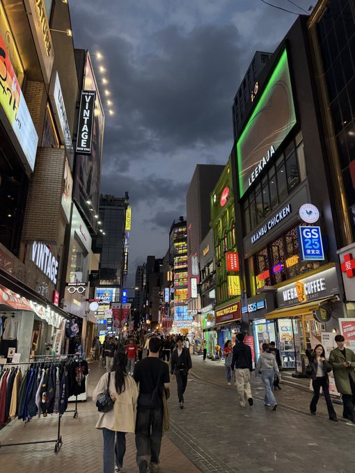 A bustling street scene at dusk in what appears to be a Japanese shopping district, featuring neon-lit signs for Vintage, LeBaja, Keimu Chicken, and Mer Coffee with pedestrians walking along the street.