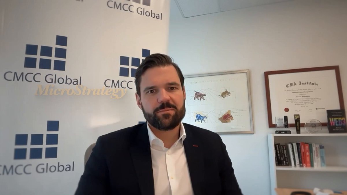 The image features a man wearing a suit sitting at a desk in front of a bookshelf filled with various items, including several books. The man appears to be posing for a photo or participating in an online meeting. The backdrop includes a sign that reads "CMCC Global" and another sign that says "Microeconomics".