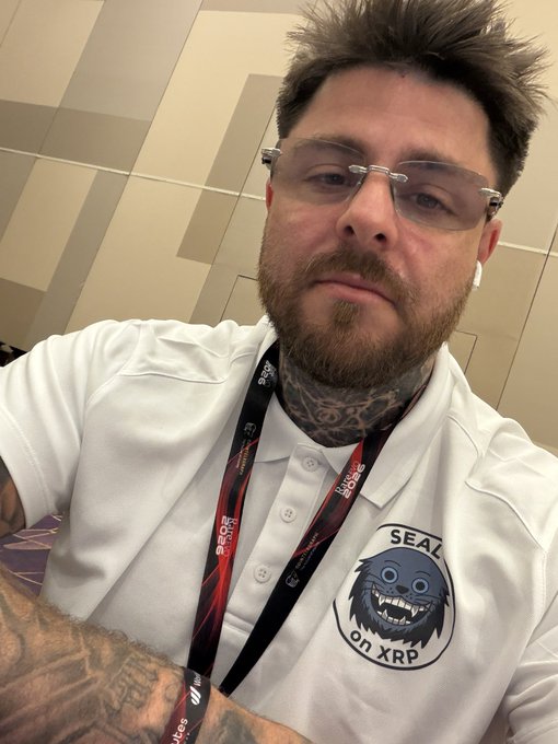A man with spiky hair, glasses, and a beard wearing a white polo shirt featuring a 'SEAL on XRP' logo and an XRP-branded lanyard.
