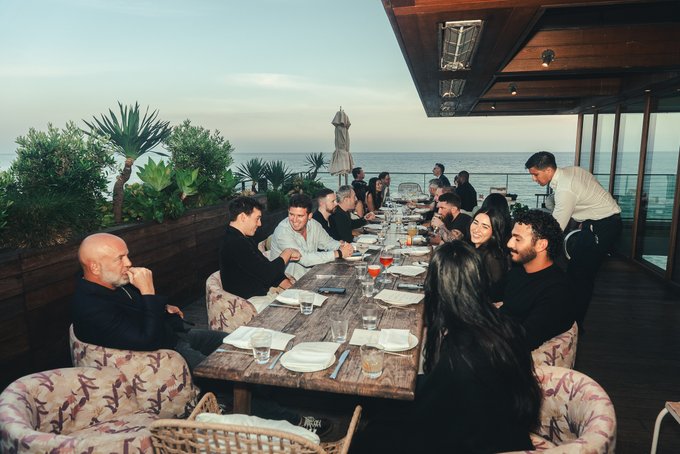 A group of well-dressed members of Introduction.com gather for an intimate dinner at an oceanfront restaurant, with a waiter serving the long table, reflecting the exclusive professional networking and business success highlighted in the accompanying post.