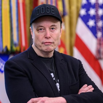 Elon Musk wearing a black "DOGE" cap stands with arms crossed in front of American flags and ceremonial banners. The image accompanies news of Musk hinting at a potential future SpaceX/Tesla merger, highlighting his expanding influence across multiple major corporations.