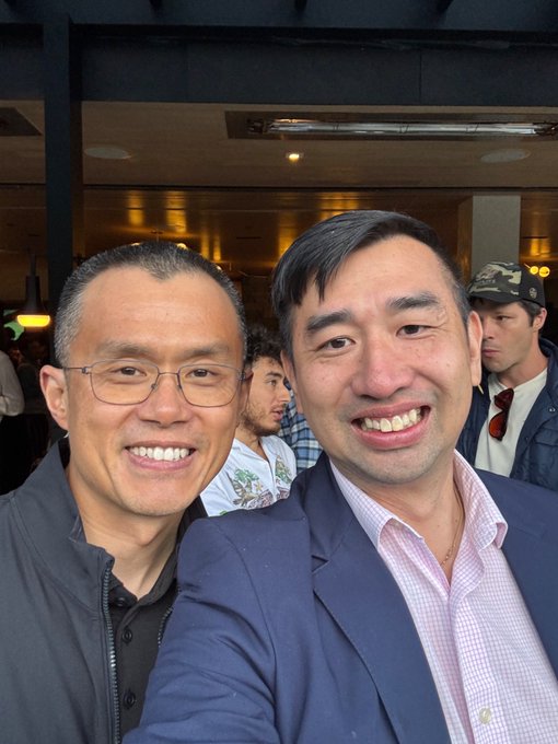 A selfie of two smiling men indoors, one wearing glasses and a grey jacket, and the other wearing a blue blazer over a pink checkered shirt.