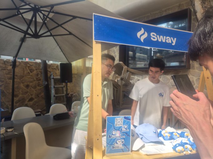 A photo of a booth at an outdoor event with a blue 'Sway' sign, where people are browsing merchandise including blue hats and t-shirts.