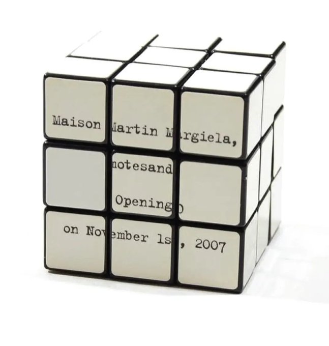 A white Rubik's cube with text on each square reading 'Maison Martin Margiela, Notes and, Opening on November 1s, 2007' across the visible faces.