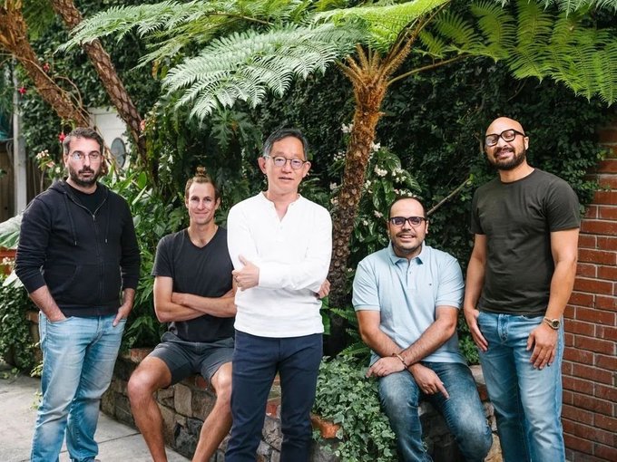 A group photo of five men posing outdoors in a garden setting with lush greenery, large tree ferns, and a brick wall in the background.