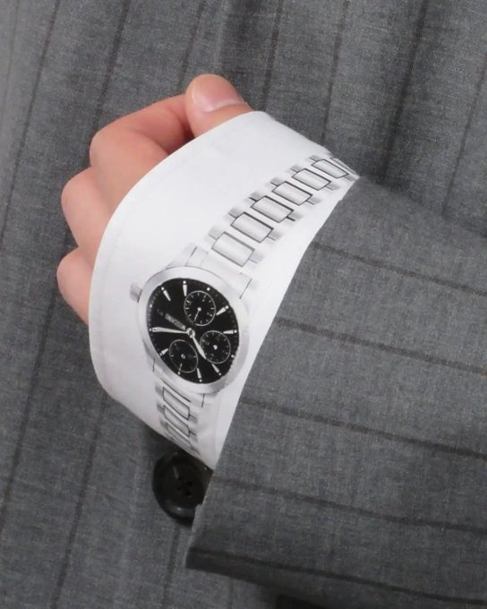 A hand holds up a silver wristwatch with a black face against a gray pinstriped suit, likely a promotional or product photo.