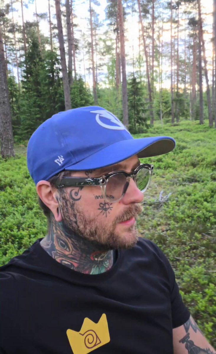 The image features a man wearing glasses, a black shirt, and a blue baseball cap. He has tattoos on his neck and face, as well as a crown-shaped yellow design on his t-shirt. The man appears to be looking off into the distance or at something in front of him. There is no text present in the image, but it seems that he might have been taking a photo with someone else who is not visible in the frame.