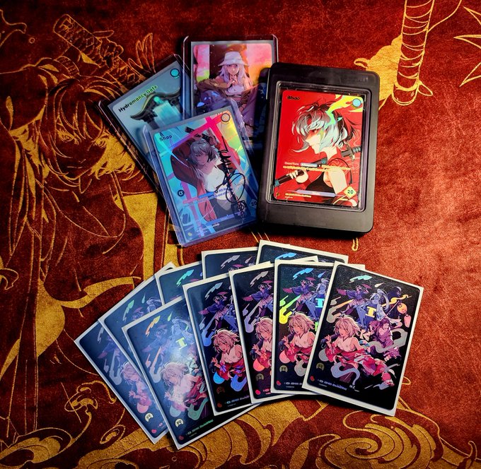 A fan spreads out their Azuki TCG holographic collectible cards and card sleeves, featuring colorful anime-style character artwork, laid across a dragon-embroidered red and gold cloth. The display showcases shiny holographic "Drop," "Once Upon," and demon-hunter themed cards with vibrant fantasy illustrations.