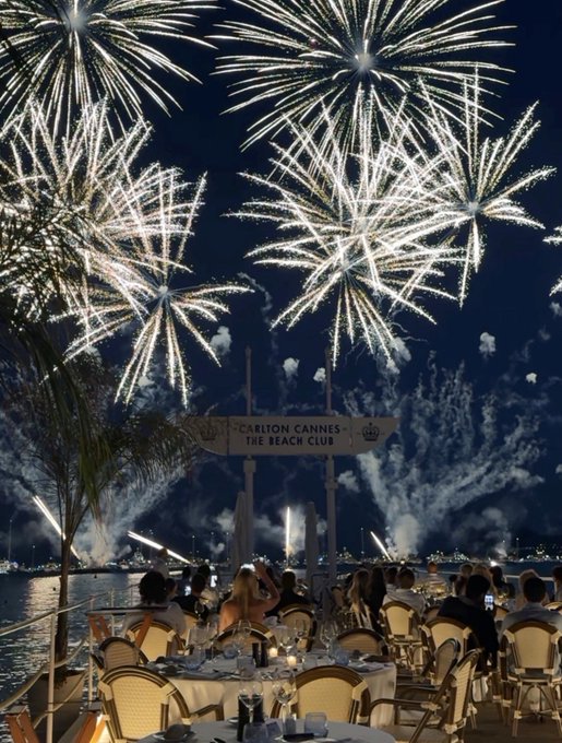 An evening scene at Carlton Cannes - The Beach Club, where diners at an outdoor restaurant are watching a spectacular fireworks display over the water.