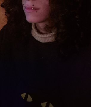 A close-up selfie of a person with curly dark hair wearing a dark sweater over a beige turtleneck, with a cat face design visible on their shirt.