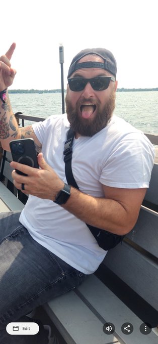 A bearded man wearing sunglasses and a backwards cap sticks his tongue out and points his index finger up while holding a smartphone, sitting on a boat on the water.