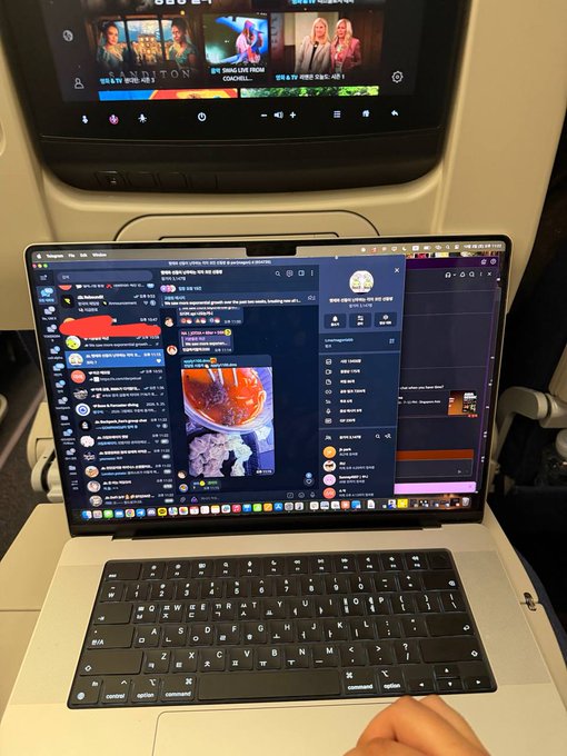A first-person perspective of someone working on a MacBook laptop during a flight, with the seatback entertainment screen visible in the background.