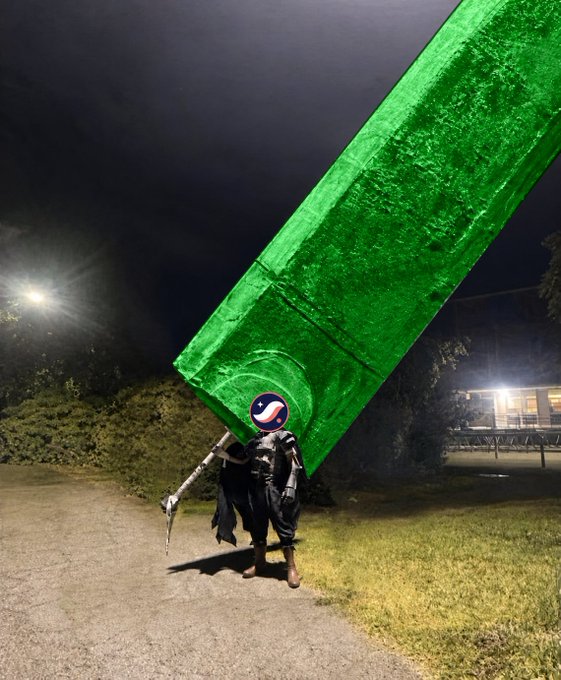 A person in a dark cloak stands on a path at night carrying a massive, oversized green beam, with a Pepsi logo sticker placed humorously over their head, against a backdrop of trees and bleachers.