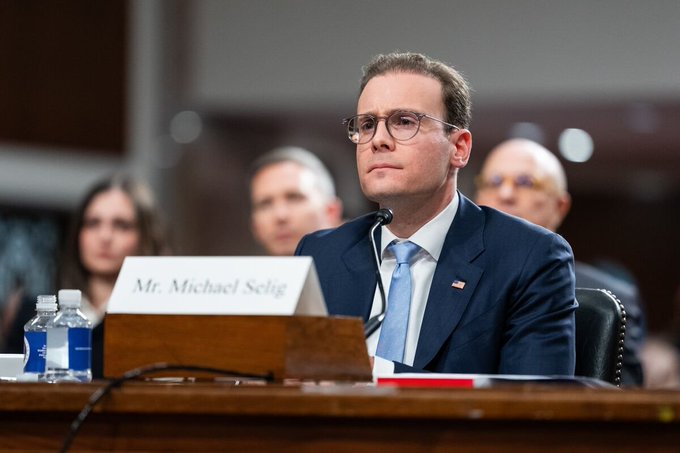 A man in a navy suit and glasses, identified by the placard as "Mr. Michael Selig," appears to be testifying or speaking at a formal hearing, with the setting and his serious expression suggesting the context of regulatory scrutiny referenced in the accompanying post about the CFTC's investigation into Polymarket.