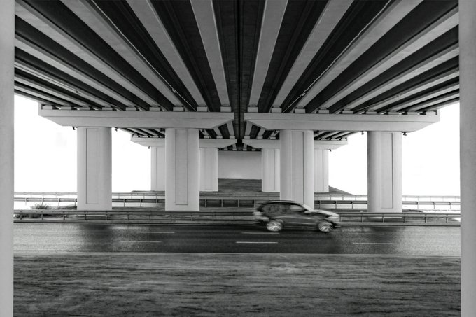 A black-and-white photograph taken from beneath a concrete bridge, showing massive pillars supporting the structure while a car drives past on the road.