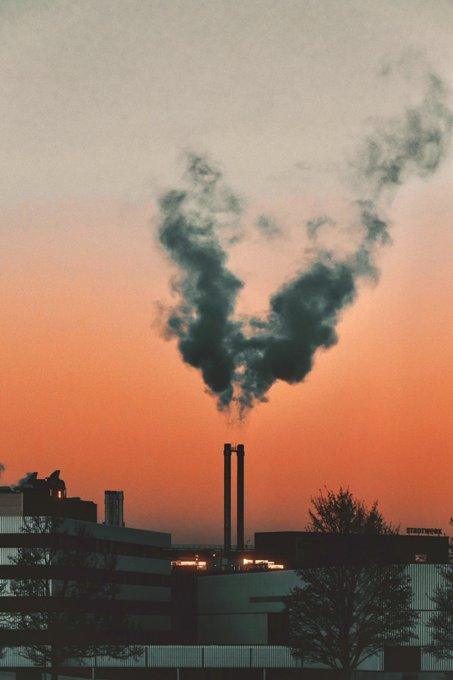 A photograph of industrial smokestacks emitting dark smoke that forms a V-shape against a vibrant orange sunset sky, with silhouetted buildings and trees in the foreground.