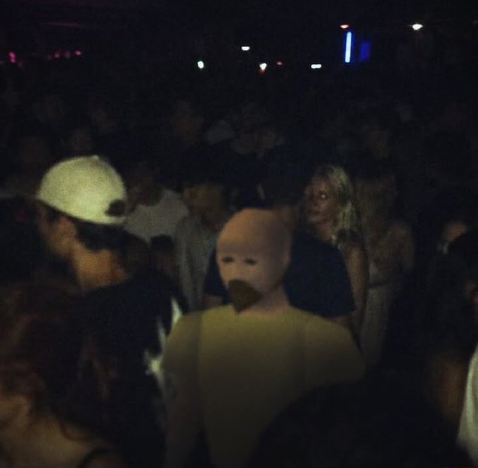 A blurry photo of a crowded nightclub scene, where a person wearing a disturbing baby mask as a beanie stands out among the dancing crowd, capturing the chaotic and surreal vibe of a late-night party.