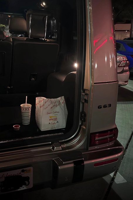 A nighttime photo of the open rear hatch of a Mercedes G63 SUV with takeout food (a drink cup and paper bag) placed on the cargo area, parked next to other vehicles.