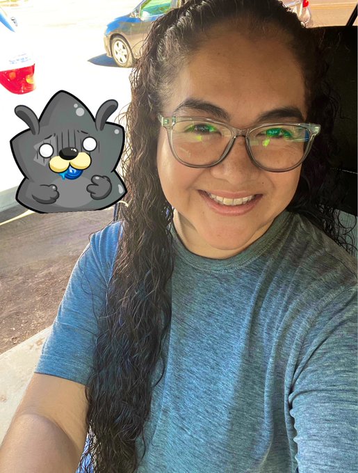 The image features a woman with long hair wearing glasses, smiling at the camera while posing for a photo. The background of the picture includes cars and buildings, suggesting that they are taking place outdoors or in an urban setting. The caption of the image reads "Summer has begun! time to enjoy with the family and creating with the most amazing family: @Kind_HQ @RoninNetwork happy #RoninMondaySelfie @KanstarWorld @AxieCreator @AxieSelfie".