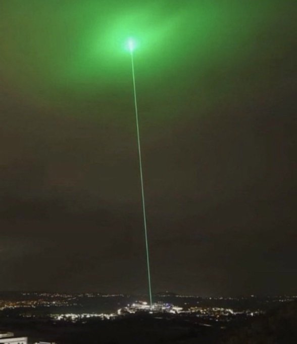 A bright green laser beam shoots up from a city at night into the dark sky, creating a glowing green cloud at its peak.