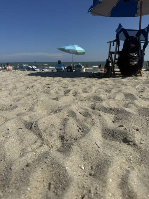 A sunny beach scene showing sand in the foreground with beachgoers, a striped umbrella, and a lifeguard chair in the distance under a clear blue sky.