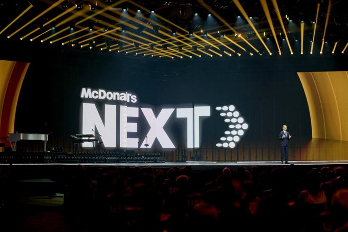 The image features a stage where a man is giving a speech at a McDonald's event. The stage has a large screen displaying the McDonald's logo, indicating that this could be an advertisement or promotional event for the fast-food chain. The audience consists of several people sitting in chairs and watching the speaker on stage. 

The background shows a black curtain with yellow lights behind it, creating a dramatic effect as if the stage is located inside a theater. There are also some hand-breaded wings visible near the center of the image, suggesting that this event might be related to the McDonald's "Next" strategy or an upcoming product launch.