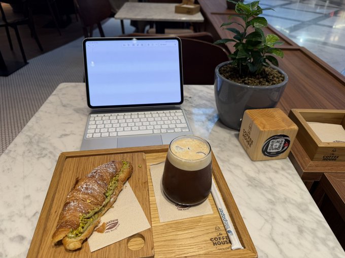 A marble café table with a laptop open to a document, a croissant sandwich, an iced coffee in a glass, and branded napkins from 'COFFEE HOUSE'.
