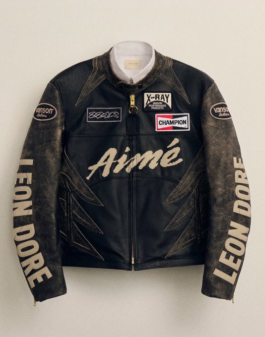 A black leather jacket with "Aimé" written in gold script across the chest, featuring "LEON DORE" lettering on both sleeves and patches including "Champion", "X-Ray Vision", and "Vanson Leathers".