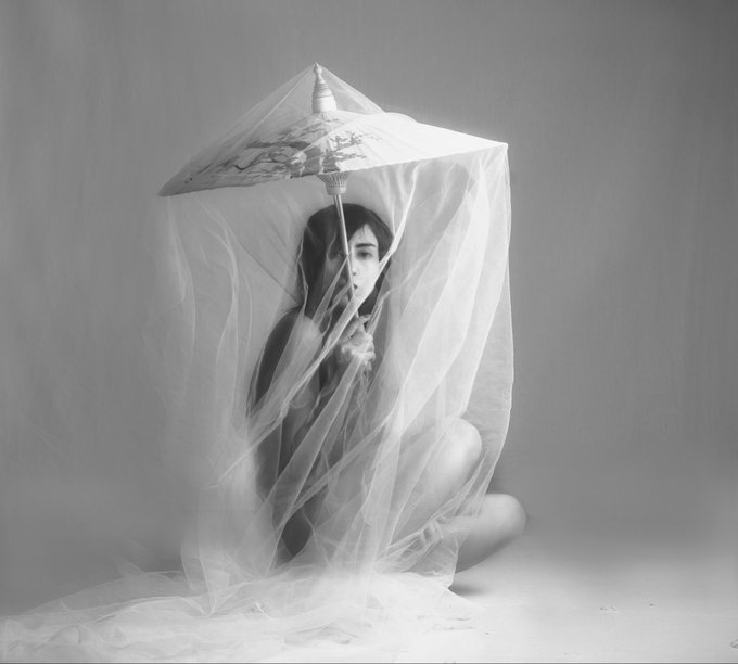 A black and white artistic photograph shows a woman sitting on the floor draped in a sheer white veil, holding a decorative parasol over her head.