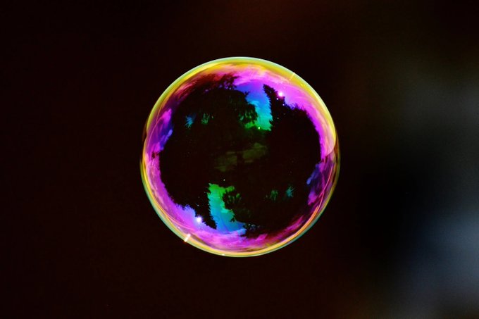 A soap bubble floats against a dark background, its surface displaying iridescent patches of pink, green, and blue.
