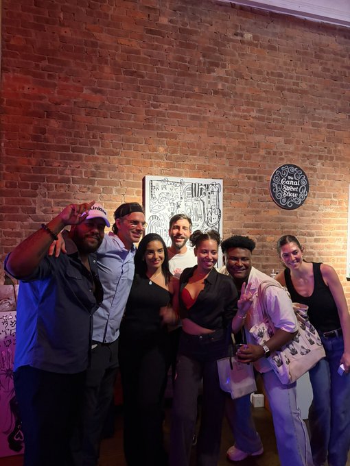 A group photo of seven people posing together indoors at what appears to be the Canal Street Station, with exposed brick walls and artwork visible in the background.