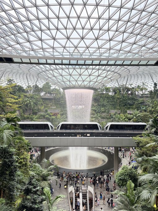 An interior view of the Jewel Changi Airport, showcasing the iconic Rain Vortex waterfall surrounded by lush greenery and the geometric glass dome ceiling.