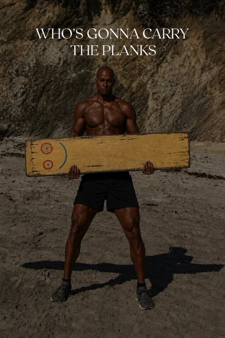 A muscular shirtless Black man standing on a sandy beach holding a yellow wooden plank with a smiley face drawn on it, with the text 'WHO'S GONNA CARRY THE PLANKS' above him.