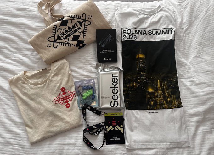 Solana Summit 2026 merchandise laid out on a white bed, including tote bags, t-shirts, a 'Seeker' phone case and device box, lanyard, and VIP contributor badge.