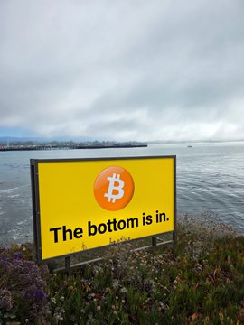 A yellow billboard with the Bitcoin logo stands along the Santa Cruz coastline, displaying the optimistic message "The bottom is in." to fellow crypto enthusiasts.