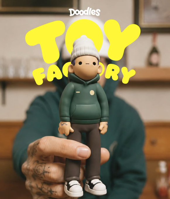 A toy figurine of a person in a beanie and green hoodie being held up by a tattooed hand, with bold yellow text reading 'Doodles TOY FACTORY' overlaid.