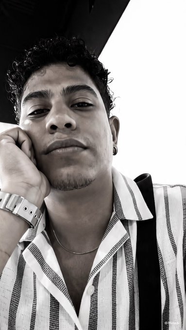 A black-and-white selfie of a young man with curly hair, wearing a striped shirt and silver watch, resting his chin on his hand.
