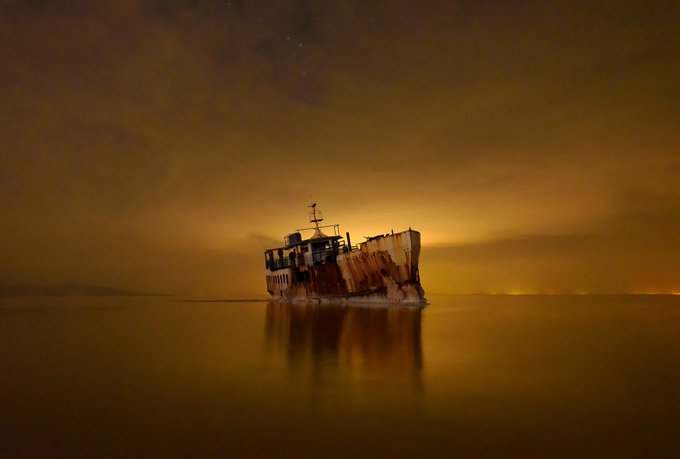 A shipwreck stands in calm waters under a moody, orange-tinted sky at night, its silhouette reflected on the still sea surface.