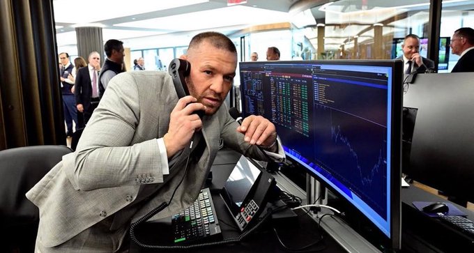 A bald man in a gray suit jacket leans over a desk covered in multiple phones, intently holding one receiver to his ear while staring anxiously at a monitor displaying stock/crypto charts with green and red tickers and a downward-trending line graph. The image humorously captures the frantic, desperate energy of someone who just made a risky meme coin purchase, with the trader surrounded by communication devices as if trying to coordinate a bailout on collapsing charts.