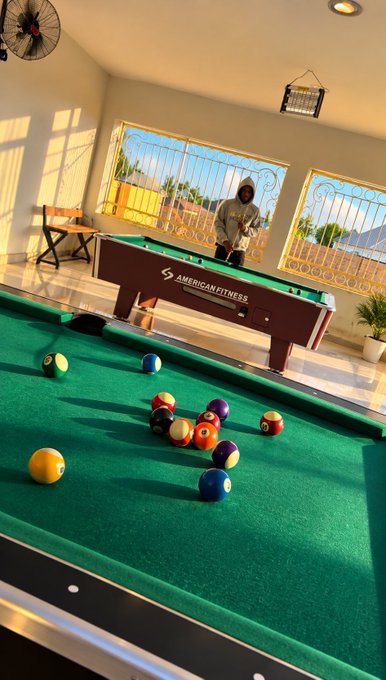 An indoor billiards room with green felt pool tables scattered with colorful balls, featuring "American Fitness" branding and large windows letting in sunlight.
