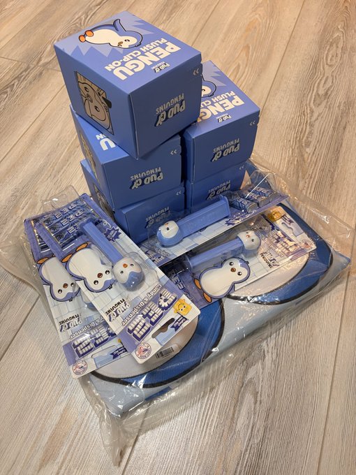 A pile of blue Pop Mart 'PENGU' blind boxes and matching plush clip-on accessories in their plastic packaging resting on a wooden floor.