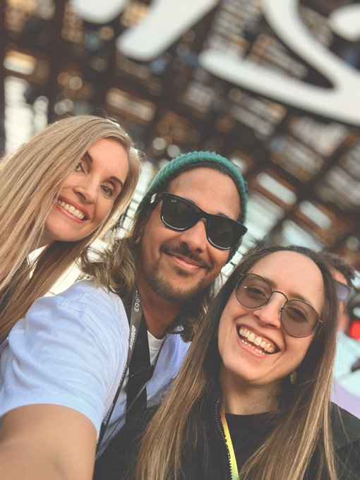Three friends smile for a group selfie, with one wearing a lanyard and a teal beanie, set against a geometric architectural backdrop—all sharing a cheerful "gm" (good morning) greeting with the crypto community.