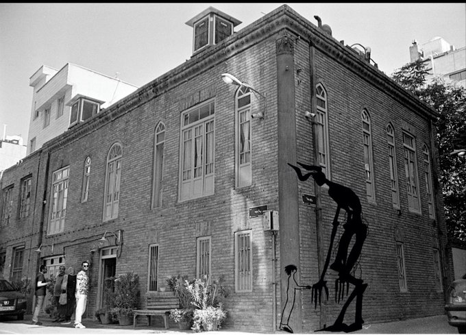 A black-and-white street view of a brick building corner featuring a large silhouette mural of a tall figure reaching toward a smaller figure on the wall, with people standing on the sidewalk below.
