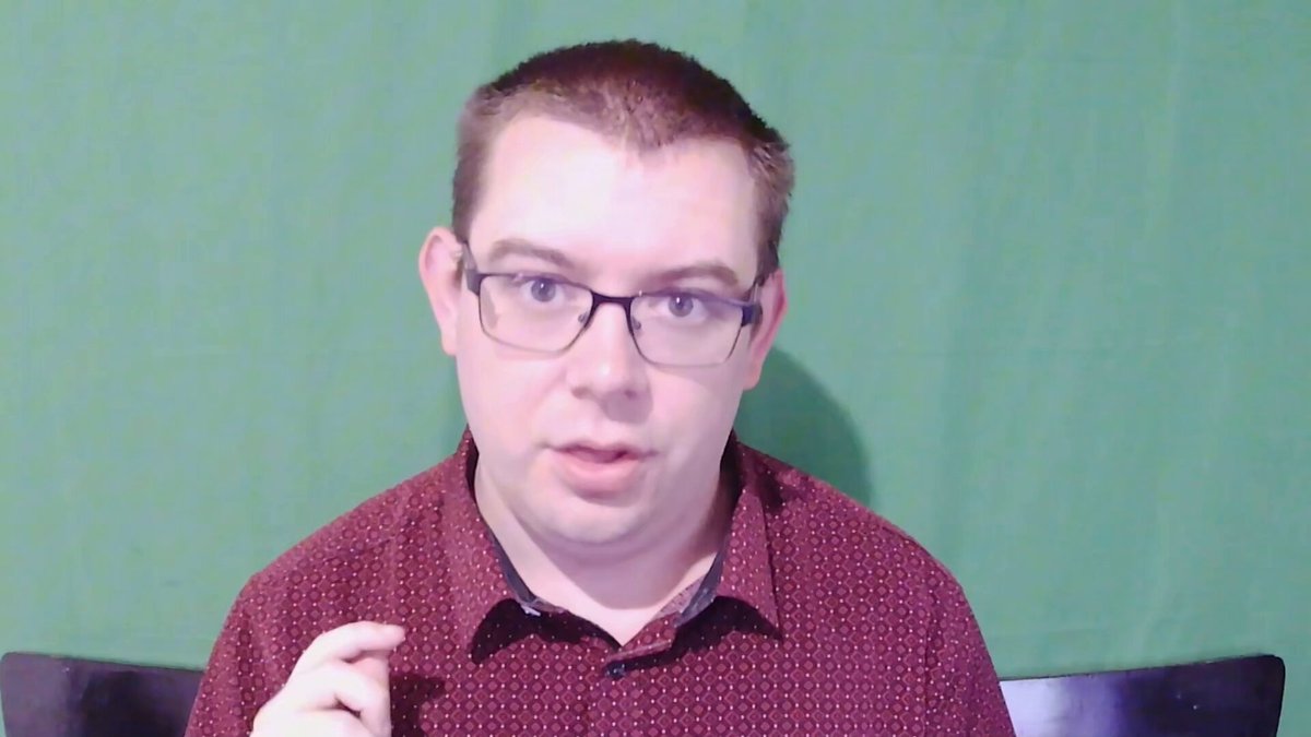The image shows a man wearing glasses sitting at a desk with his hands up in front of him. The background appears to be green, which could possibly be a computer screen or a large window. There are no other people visible in the photo. The main focus is on the man and his actions, as he seems to be making a gesture or performing an action that might be related to the post about the Jackpot victory.