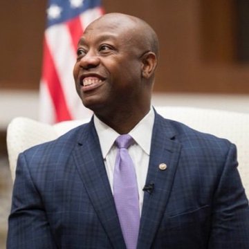 Senator Tim Scott is shown smiling warmly, wearing a dark navy pinstripe suit with a white shirt and lavender tie, seated in what appears to be an official setting with an American flag visible behind him. The image aligns with the post highlighting his push for a timely Senate vote on the Clarity Act, reinforcing his call for swift legislative action.