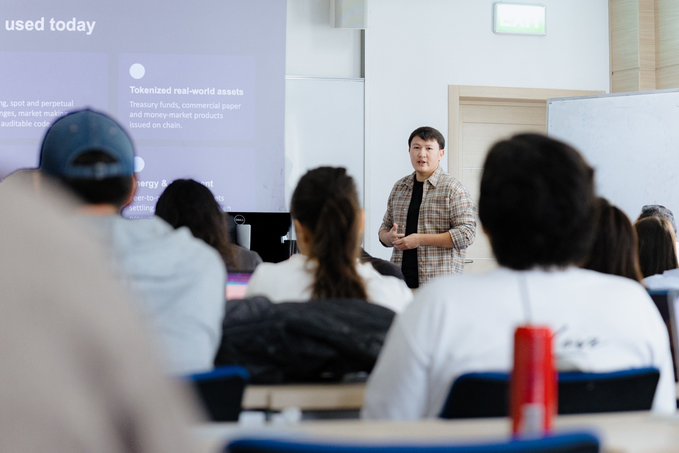 An instructor stands in front of a classroom delivering a presentation on a projection screen that reads 'Tokenized real-world assets,' discussing treasury funds, commercial paper, and money-market products on chain.