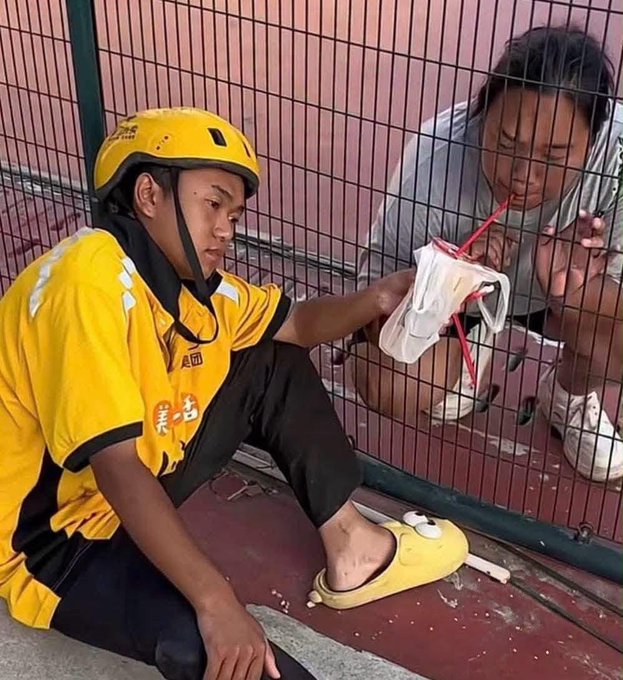 A Meituan delivery driver wearing a yellow helmet and jersey sits on the ground by a metal fence, handing a packaged food item through the fence to a woman on the other side.