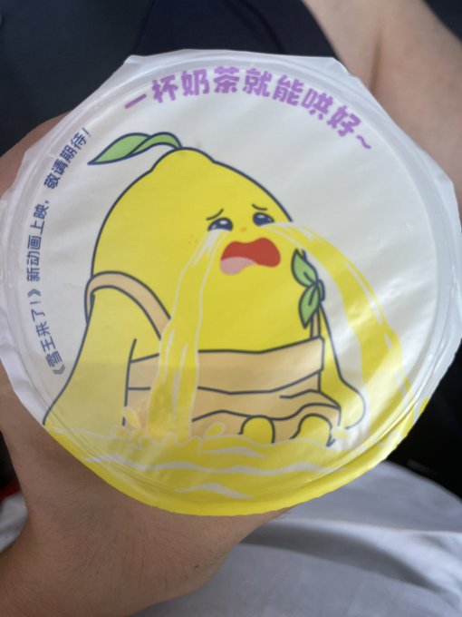 A hand holding a sealed cup of bubble tea featuring a sad, crying cartoon lemon character on the lid, with Chinese text reading '一杯奶茶就能哄好~' (One cup of milk tea can cheer me up~).