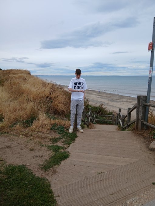 A person wearing a 'NEVER GOON' t-shirt and gray sweatpants stands on a wooden walkway looking at their phone, with the ocean and beach in the background.