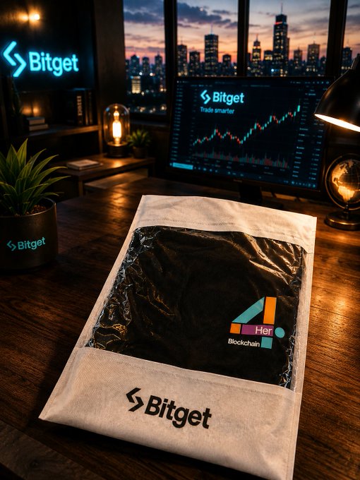 A Bitget promotional scene featuring a branded drawstring bag containing black merchandise, set on a wooden desk with a computer monitor showing Bitget's trading interface and a city skyline at dusk in the background.